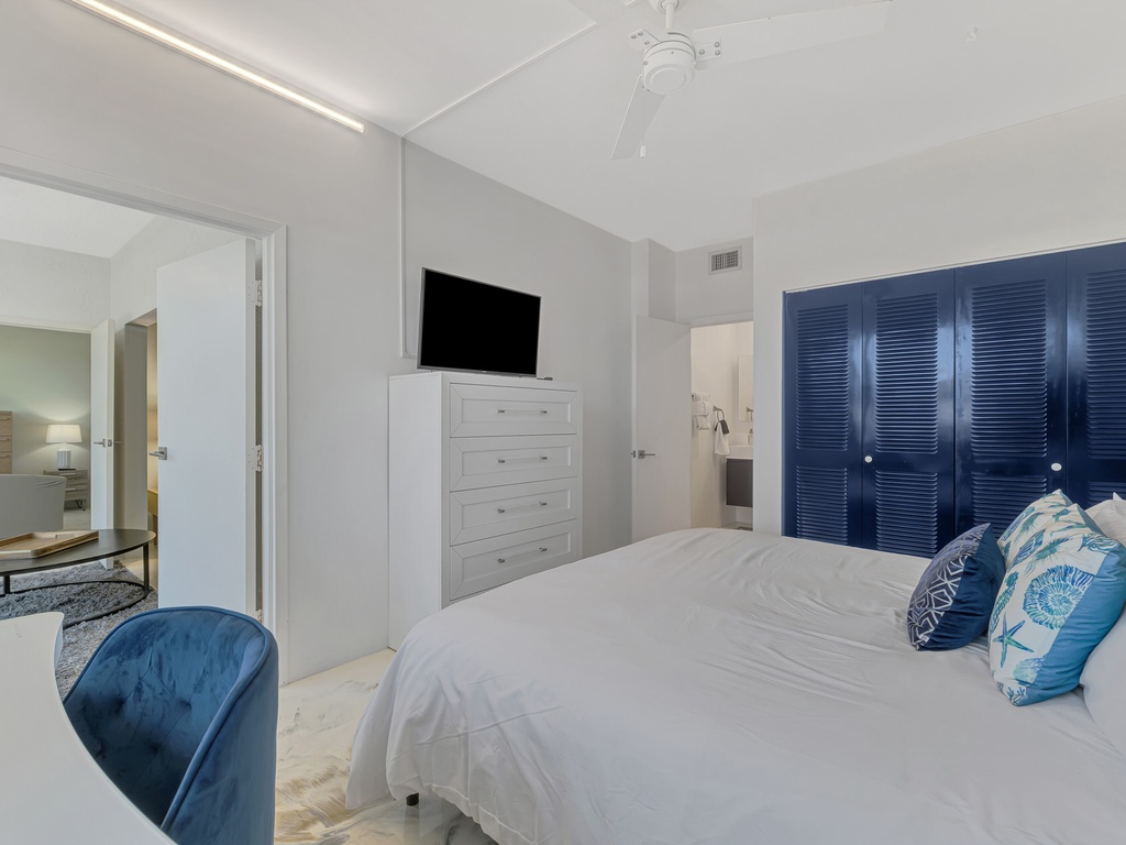 Drift off in your serene bedroom featuring crisp white linens, ocean-blue accents, and modern amenities including TV and ample storage.