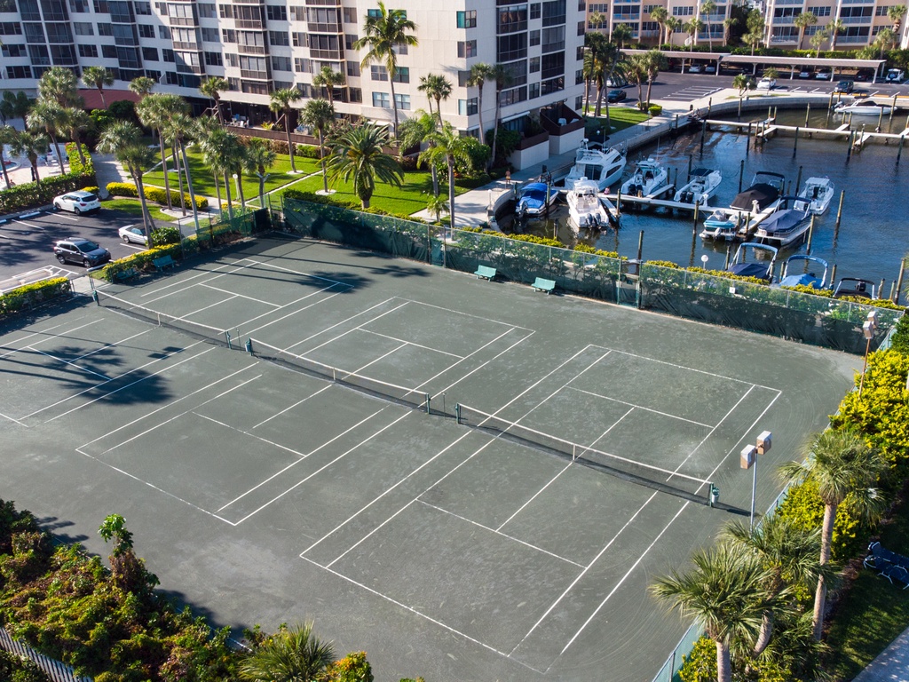 Aerial view of tennis courts surrounded by tropical landscaping with marina and residential buildings nearby.