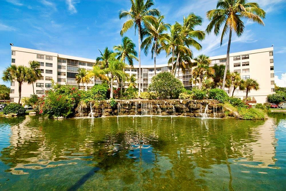 Tropical resort building with waterfront setting, featuring lush palm trees and landscaped grounds under bright blue skies.