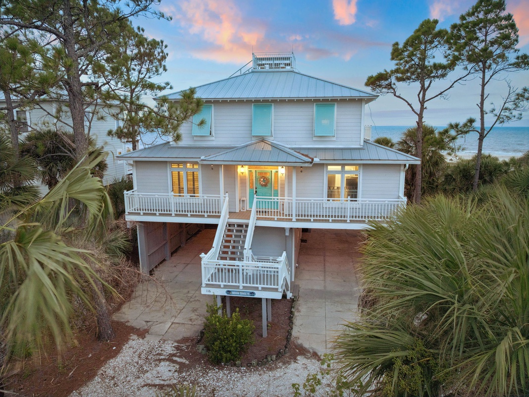 A charming coastal retreat with wraparound porches and turquoise shutters nestled among native palms and pines, just steps from the water's edge at sunset.