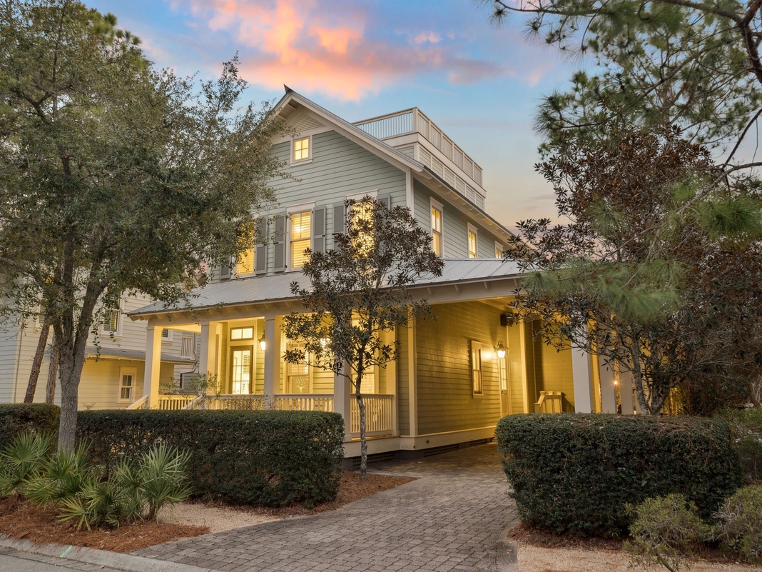 A beautiful coastal home welcomes guests with warm lighting and mature landscaping as evening approaches.