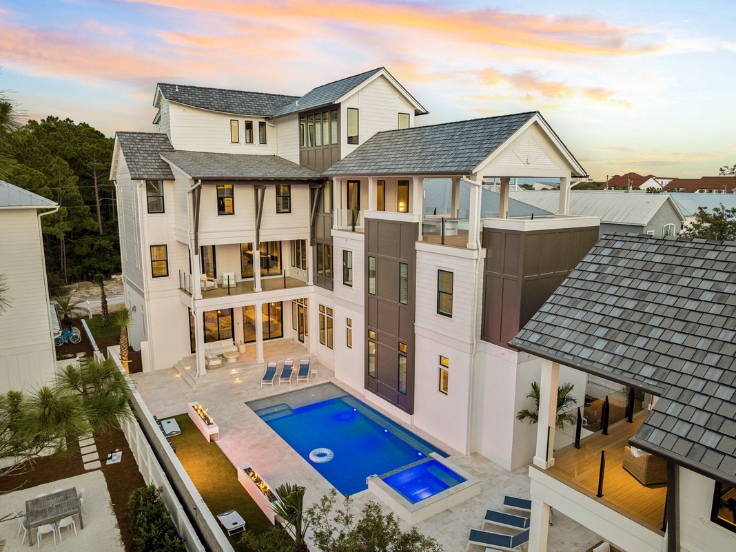 A stunning multi-level vacation home showcasing modern coastal architecture with an inviting pool and spacious outdoor areas, set against a beautiful sunset backdrop in a peaceful residential neighborhood.