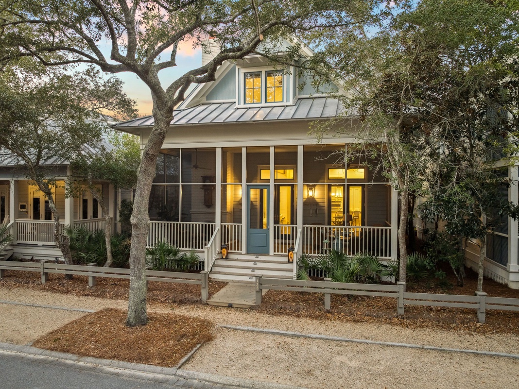 Charming cottage-style home with screened porch and metal roof nestled among mature oak trees in a peaceful neighborhood setting.
