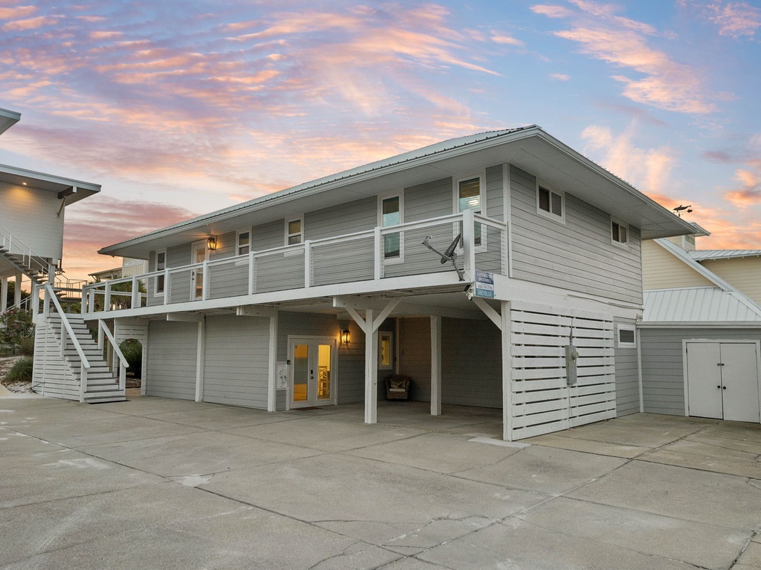 Coastal-style vacation home features stilted construction and ample parking, with sunset-painted skies above the neighboring beach community.