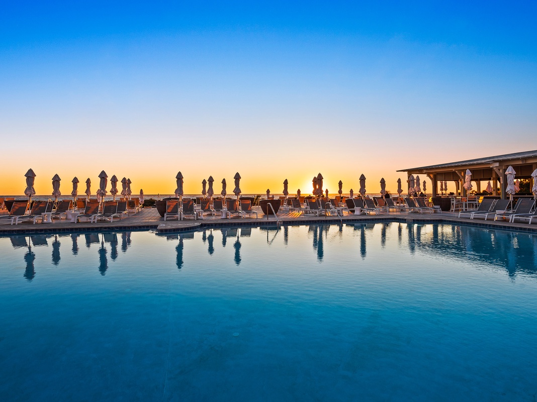 A stunning beachfront pool awaits, lined with loungers and furled umbrellas, where golden hour transforms the water into a mirror. The nearby pavilion beckons for refreshments while the horizon glows warmly.