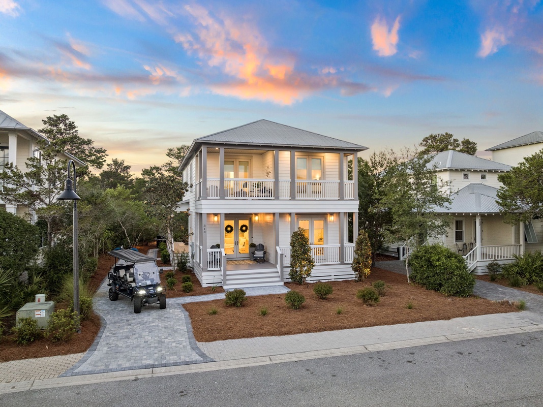 A charming two-story coastal cottage with wraparound porches and welcoming yellow doors, nestled in a peaceful residential neighborhood with paver walkways and golf cart access.