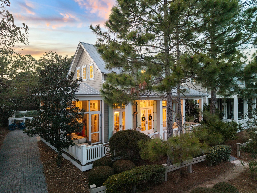 Charming coastal cottage with warm lighting glowing through French doors at dusk, surrounded by mature trees and manicured gardens.