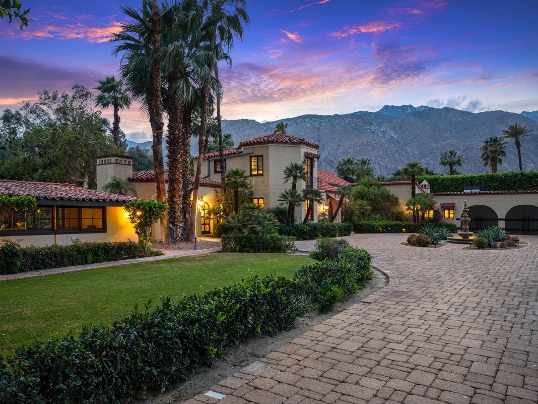 Spanish-style estate nestled against dramatic mountain backdrop, surrounded by lush palms and landscaped grounds, showcasing a welcoming entrance at twilight.