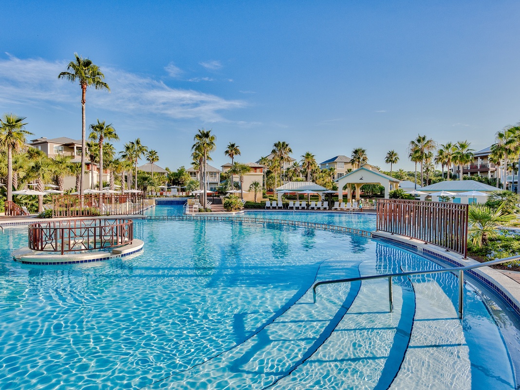 Resort swimming pool with palm trees and tropical buildings under clear blue skies.