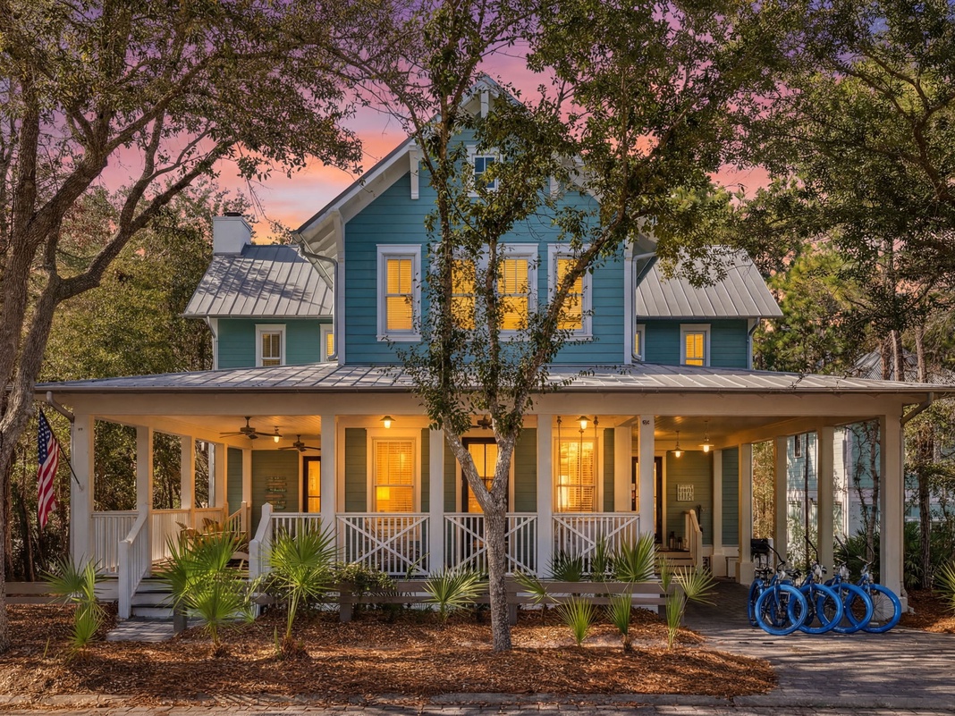 Charming blue cottage nestled among oak trees with wraparound porch and bicycles ready for coastal adventures at sunset.