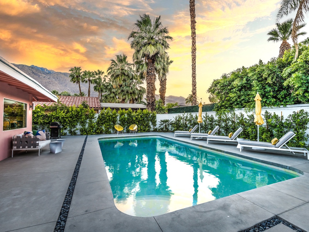 A stunning desert oasis set against dramatic mountain scenery. Your private pool sparkles under golden hour skies, surrounded by swaying palms and modern lounge chairs—perfect for soaking up the California sunshine.
