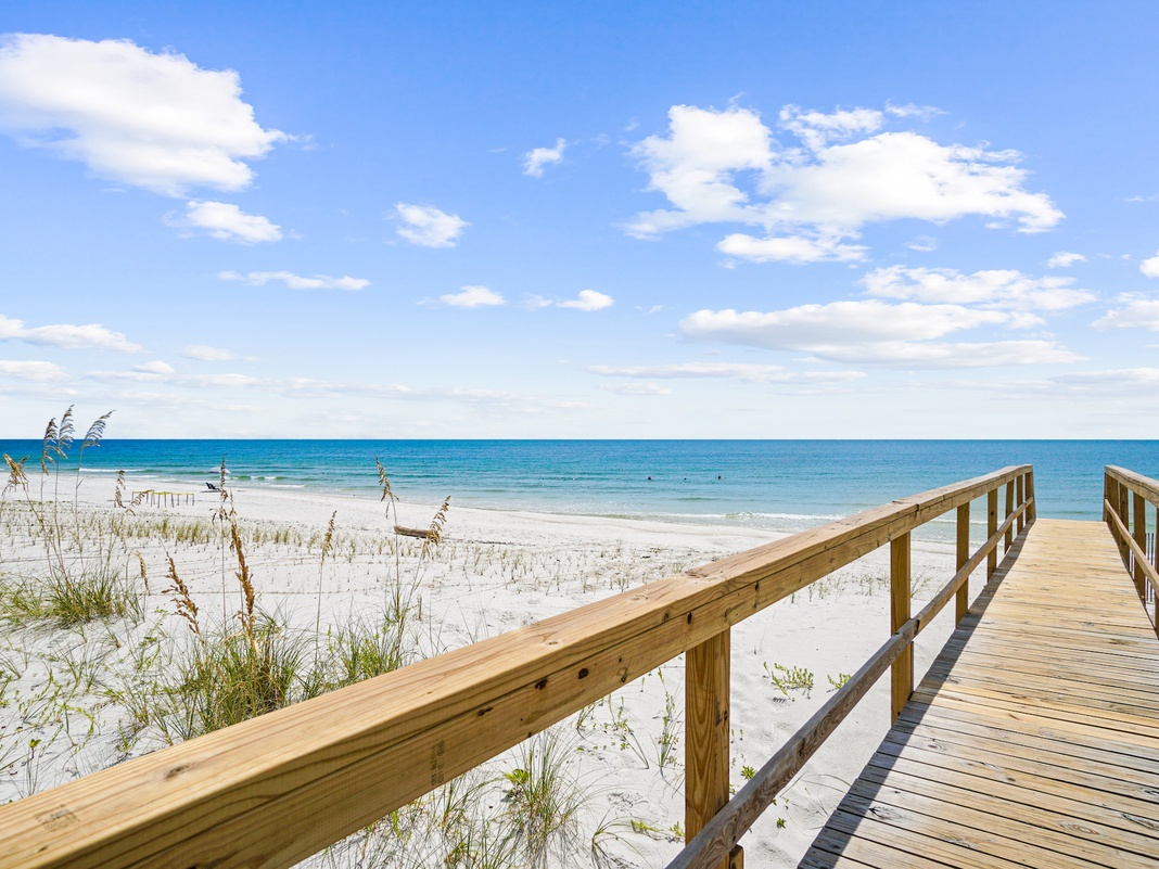 Wooden boardwalk leads to pristine white sand beach with crystal-clear turquoise waters under sunny blue skies.