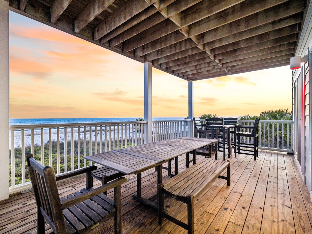A covered oceanfront deck offers outdoor dining beneath striking architectural beams, with a tranquil beachfront and sunset views as your backdrop.