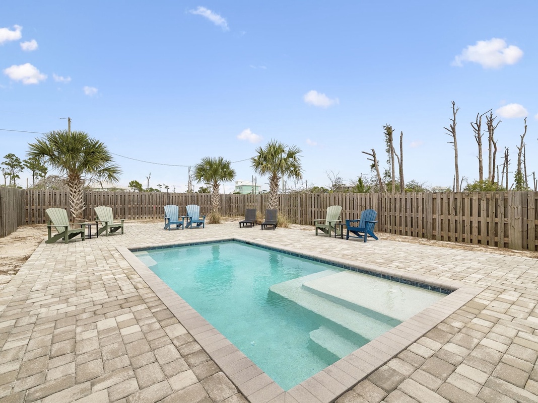 A private pool surrounded by paver stones and colorful Adirondack chairs awaits beneath clear blue skies, framed by palm trees and a secure privacy fence.