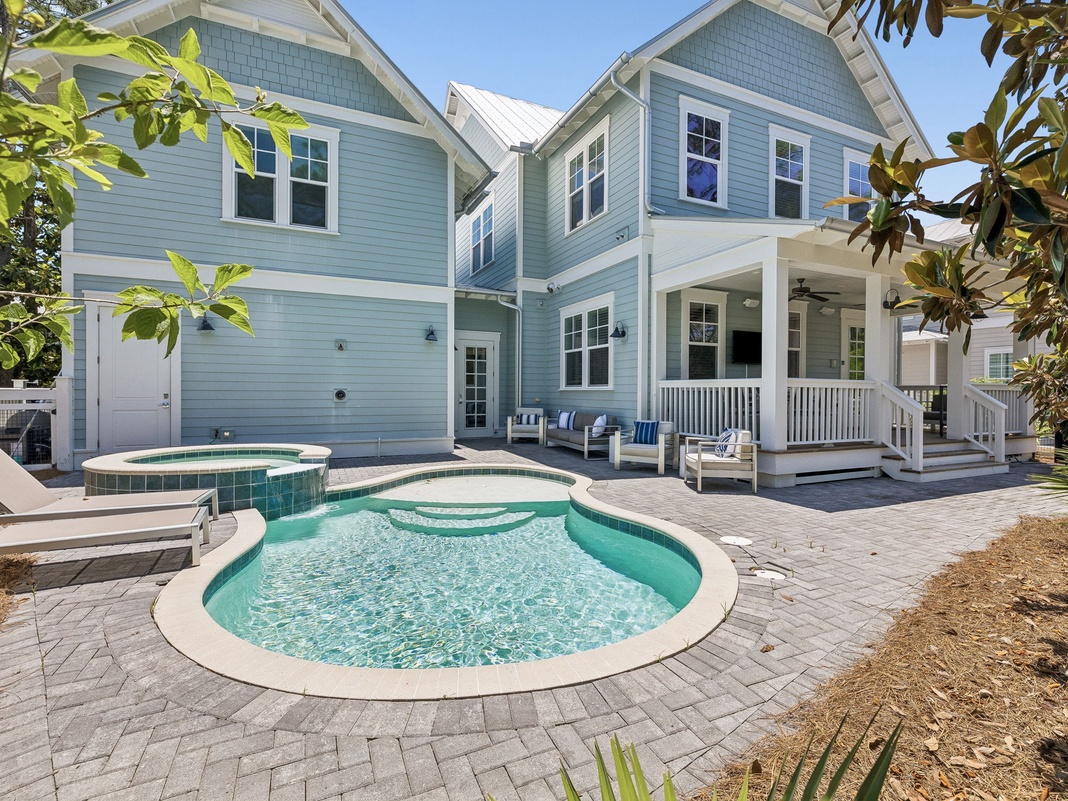 This coastal-style home features a sparkling pool, hot tub, and covered porch with ample seating—ideal for relaxing under sunny skies surrounded by lush tropical greenery and beachside charm.