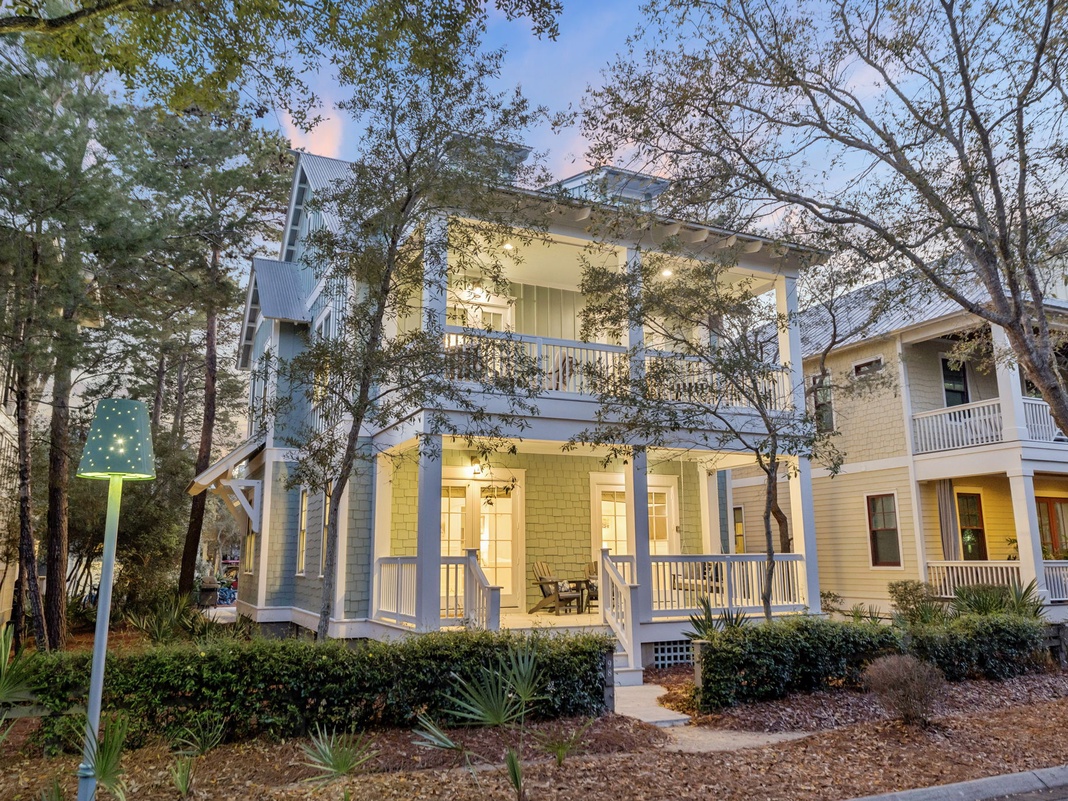 Charming coastal cottage with inviting front porches and warm lighting nestled among mature trees in a peaceful neighborhood setting.