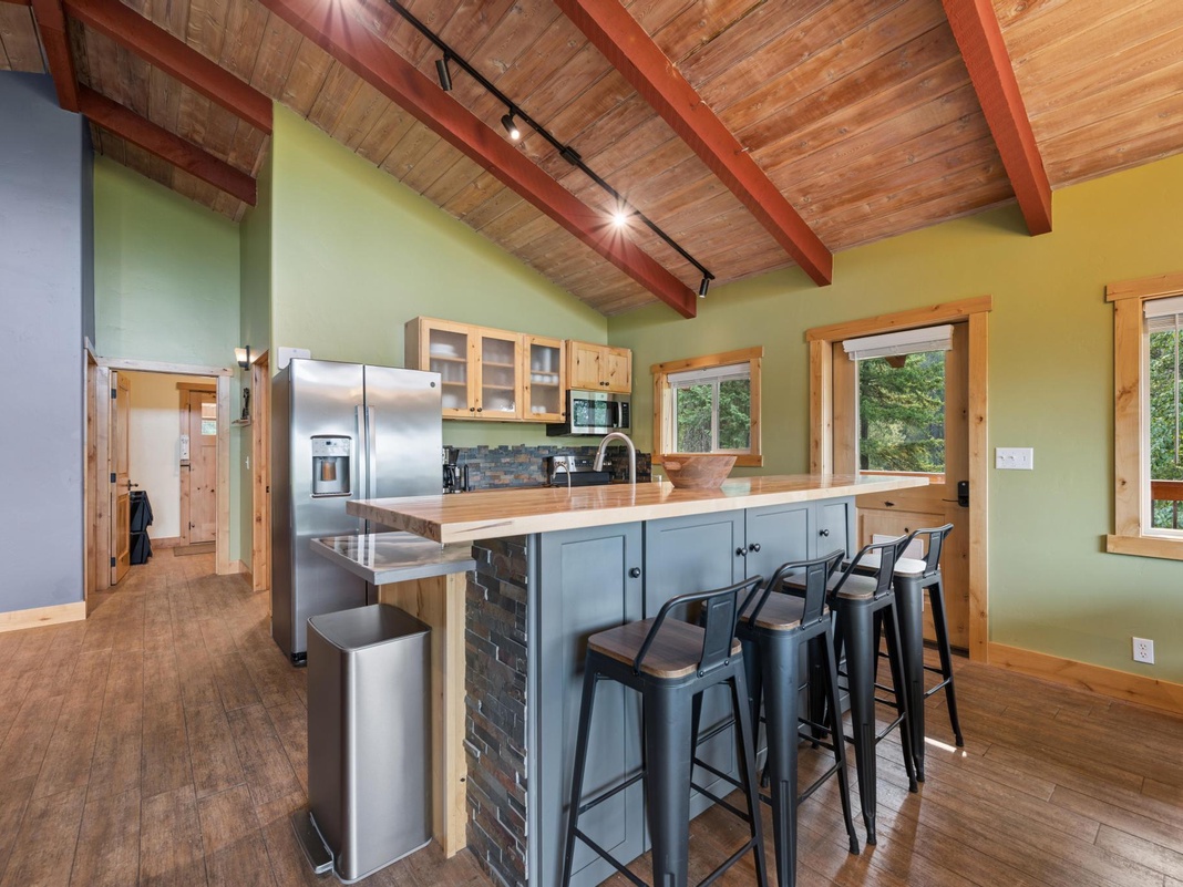 Gather around your spacious kitchen island with rich wood countertops and rustic charm. Natural light pours through generous windows, highlighting the soaring wooden ceiling beams and open-plan design perfect for shared meals and memories.