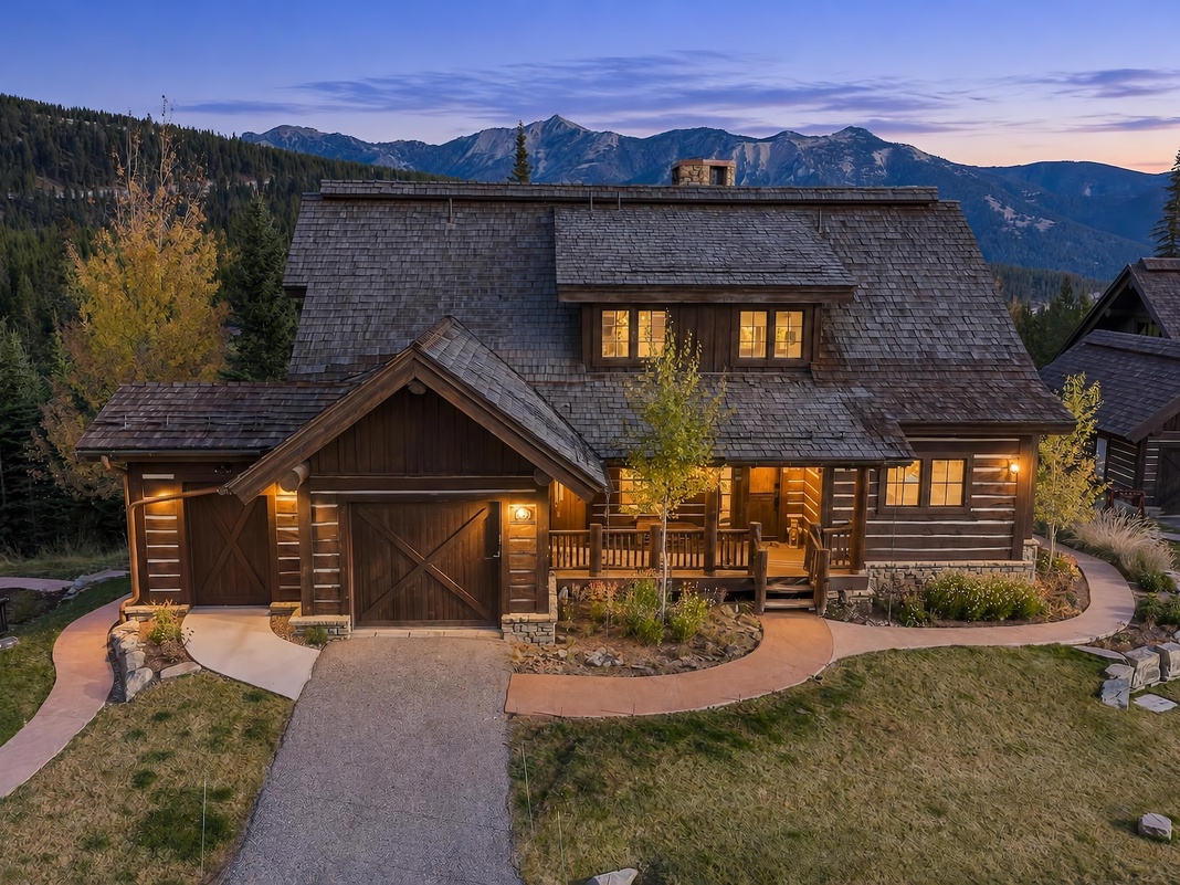 A charming log cabin nestled in the mountains, surrounded by towering pines and dramatic peaks. The warm glow from the windows and rustic wood architecture create an inviting alpine retreat as the day transitions to twilight.