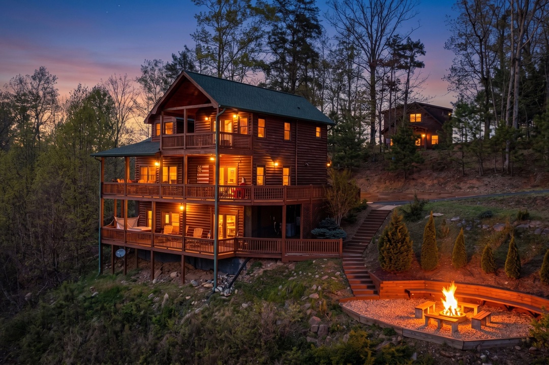 A stunning log cabin glows warmly against the twilight sky, surrounded by towering trees and natural landscape. Multiple decks and a welcoming fire pit await your mountain retreat.