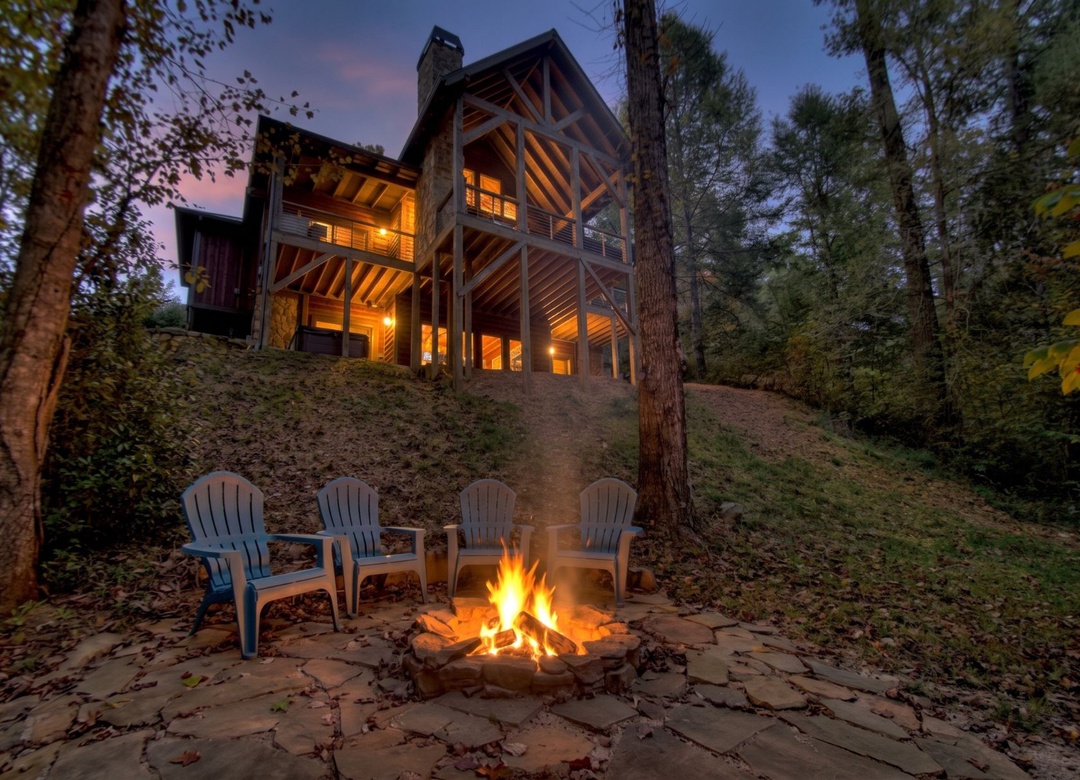 A stunning mountain cabin awaits as twilight settles over the forest, where a crackling fire pit invites you to gather beneath towering trees and glowing windows.