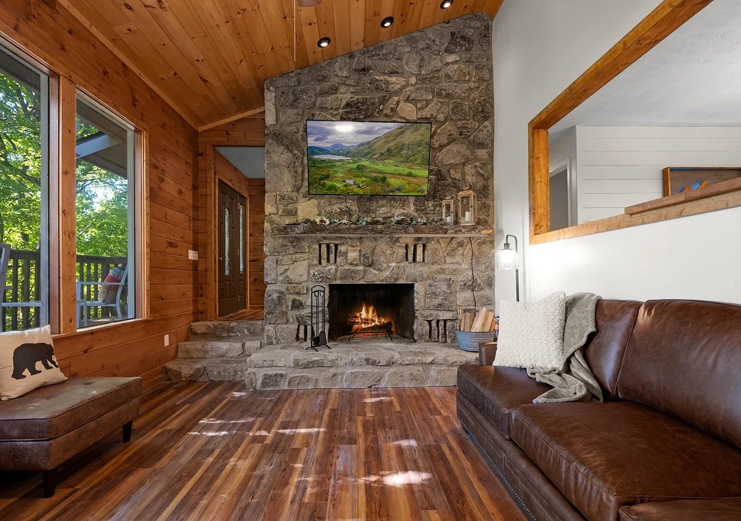 Settle into the warmth of your cabin retreat. A crackling stone fireplace invites you to unwind on plush leather seating while natural light pours through floor-to-ceiling windows, framing peaceful forest views from your private balcony.