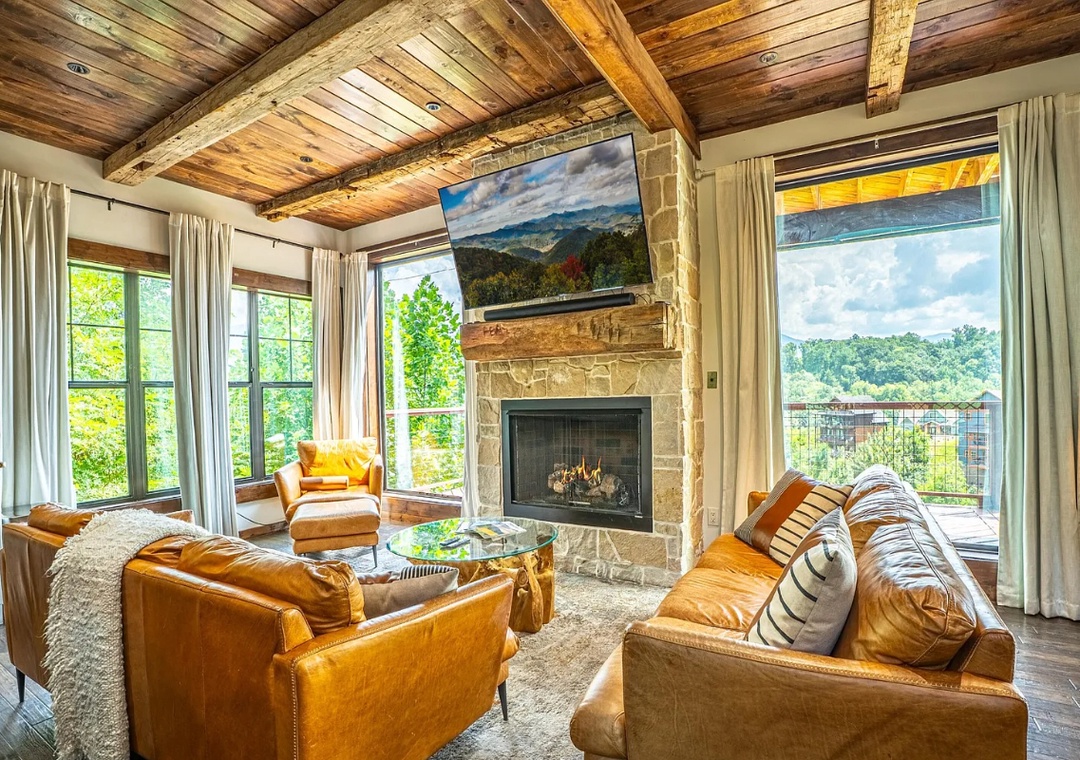 Unwind in your bright living room where rustic wood beams meet rich leather seating. Floor-to-ceiling windows frame leafy views, while the stone fireplace adds warmth and character to your mountain retreat.