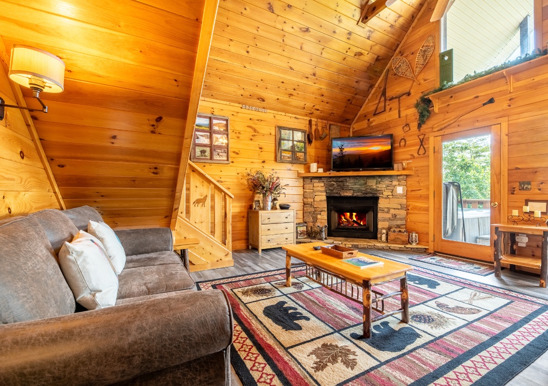 Picture yourself unwinding in this cozy cabin living room, warmed by the stone fireplace beneath soaring wood-beam ceilings, with soft seating and rustic charm all around you.