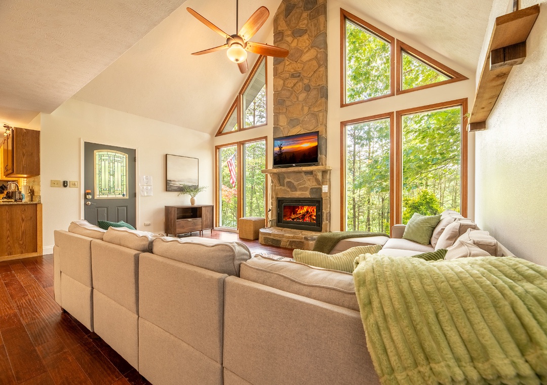 Unwind in your sunlit living room with soaring cathedral ceilings, a cozy stone fireplace, and floor-to-ceiling windows framing tranquil forest views—your perfect retreat after a day of adventure.
