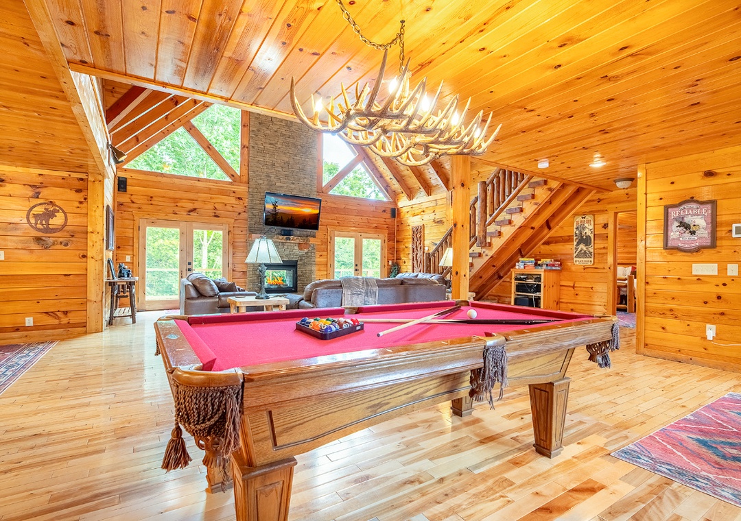 Cathedral ceilings and knotty pine walls frame this great room, featuring a pool table, stone fireplace, and cozy seating beneath an antler chandelier. Towering windows flood the space with natural light and views of the trees.