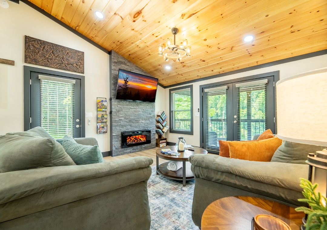 Sink into plush sofas beneath warm pine ceilings, where the electric fireplace sets a cozy mood and forest views frame your peaceful retreat through expansive windows.