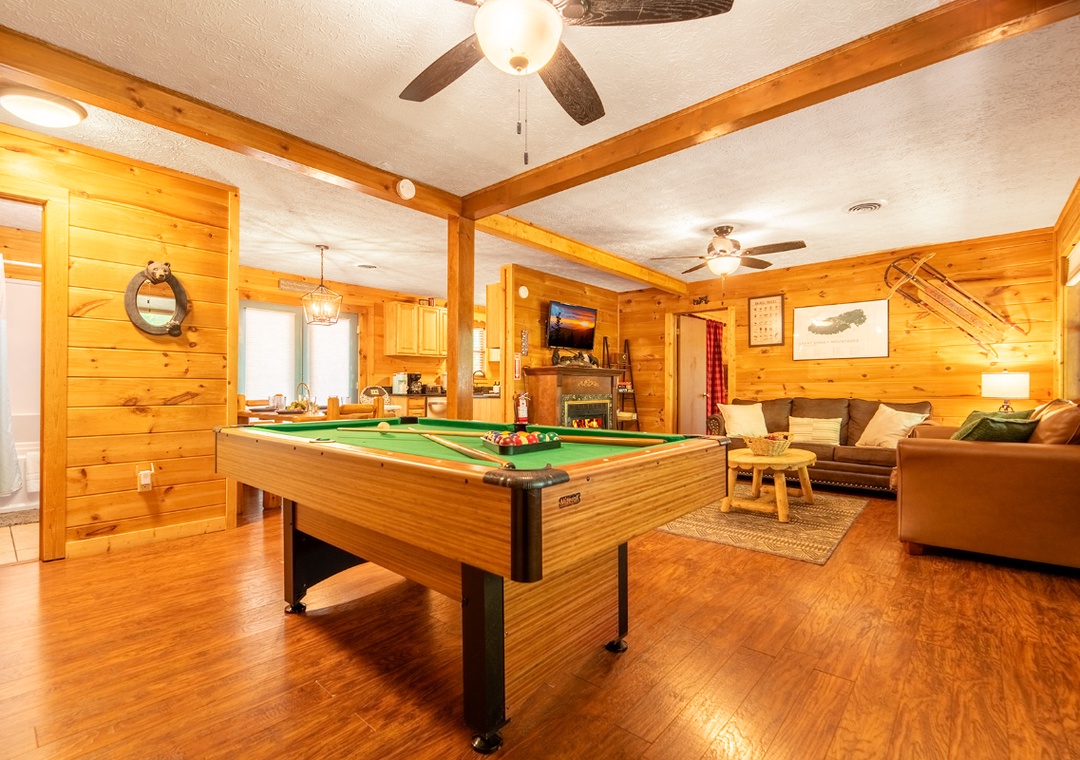 Gather around the billiard table in your cozy, open-concept game room with warm wood beams, a comfy living area, and a peek into the kitchen—perfect for friendly competition and relaxation.