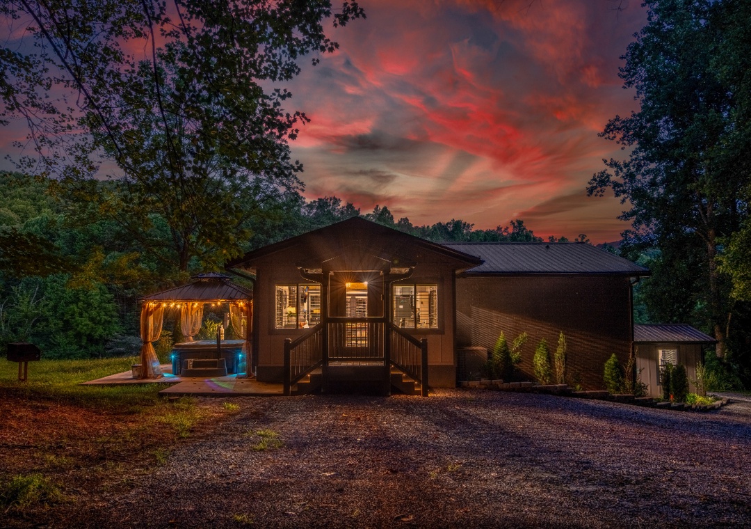 The property sits nestled among trees beneath a stunning sunset sky, with a welcoming exterior that glows warmly at dusk and a hot tub ready for relaxing evenings.