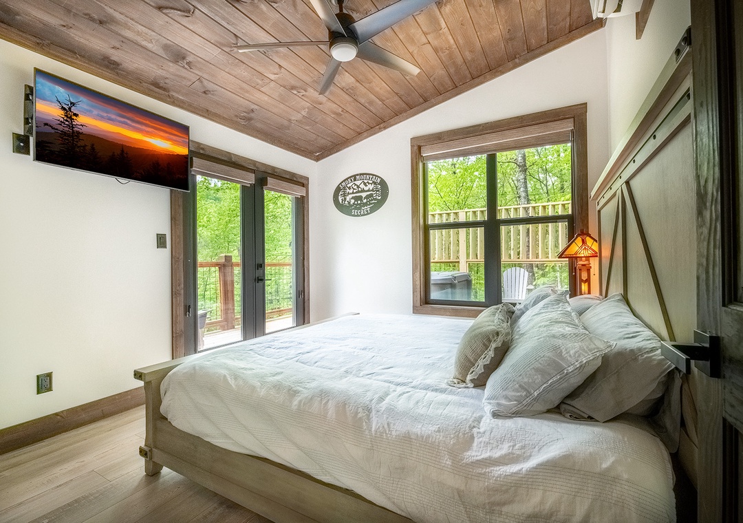 Step into your serene bedroom with rustic wood ceilings and nature-filled views. Your cozy bed awaits beneath a ceiling fan, while doors open to a private deck overlooking lush trees.