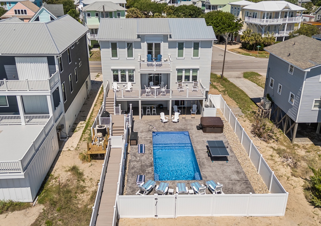 Aerial view of the vacation rental property in a coastal residential area, showcasing the private pool, hot tub, outdoor lounging spaces, and multiple surrounding beach homes.