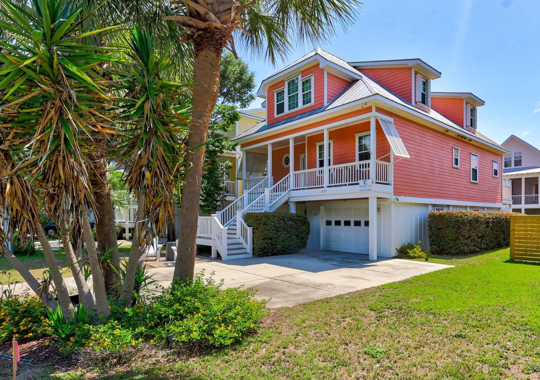 A vibrant coral-pink coastal home with welcoming covered porch, two-car garage, and tropical landscaping including mature palm trees.