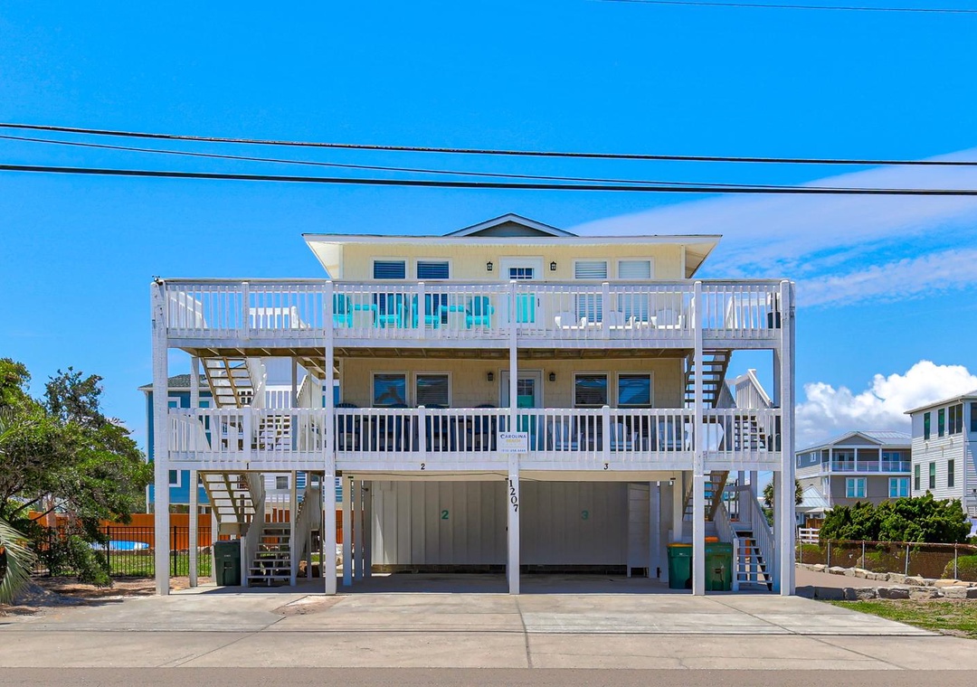 Bright coastal property with spacious balconies, elevated parking, and neighboring beach homes under a clear blue sky.