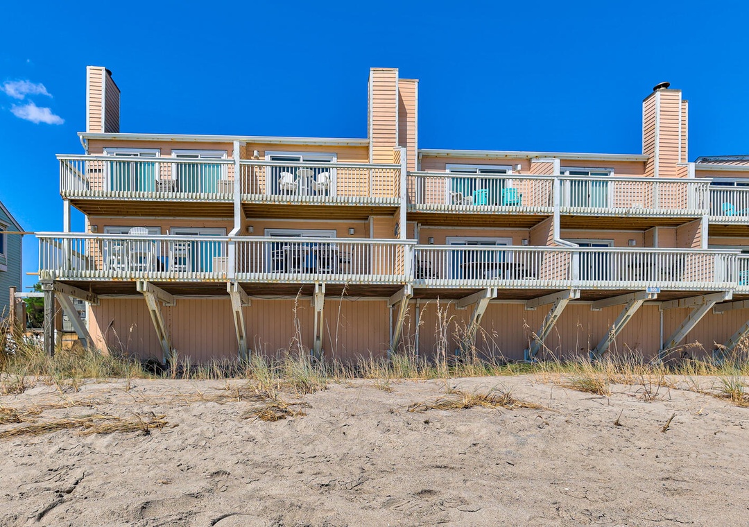 Beachfront townhomes with private balconies sit directly on sandy shores under brilliant blue skies.