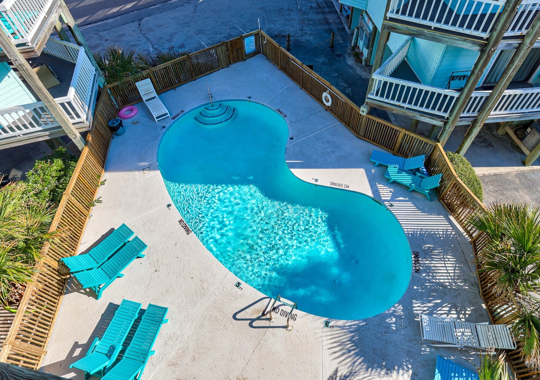 Aerial view of a sparkling blue swimming pool surrounded by tropical landscaping and comfortable lounge chairs within a gated property area.