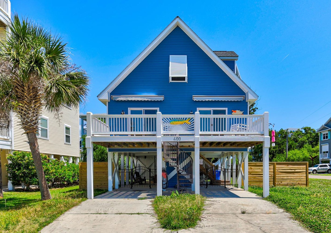 A vibrant coastal beach house elevated on stilts, featuring covered parking, a private balcony, and colorful seaside charm in a sunny residential neighborhood surrounded by palms.