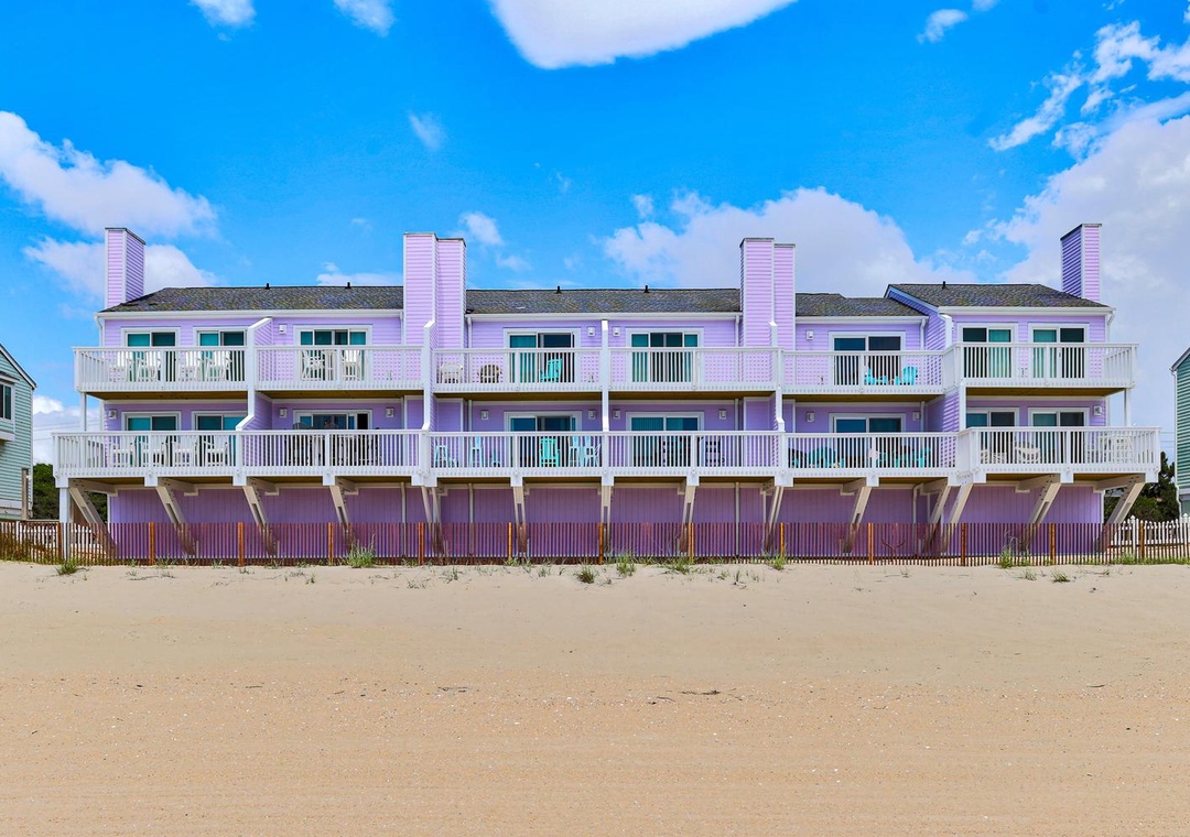 A beachfront row of vacation townhomes with multiple balconies and direct beach access under a bright blue sky. Each unit features multiple levels and private outdoor spaces.