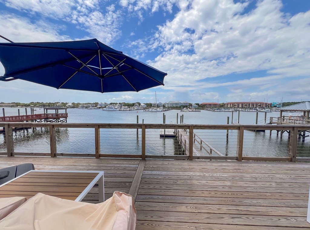 A private waterfront deck featuring sunbeds and a blue umbrella overlooks a tranquil marina with docked boats and distant coastal buildings framed by a scenic sky.