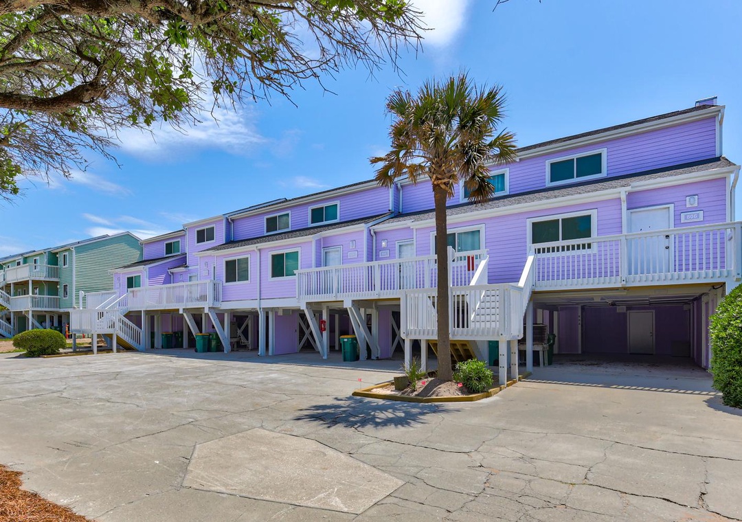 A row of colorful vacation townhomes with covered parking and palm trees under a bright blue sky.