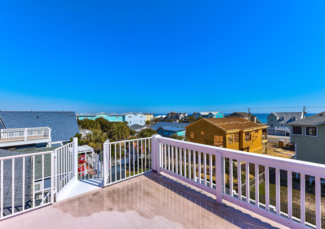 Expansive deck with white railings overlooking a charming coastal neighborhood with glimpses of the ocean beyond.