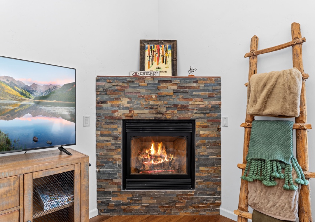 Curl up in your cozy living room with a warm fire crackling, a rustic stone fireplace, and mountain views on screen—perfect for unwinding after your adventures.