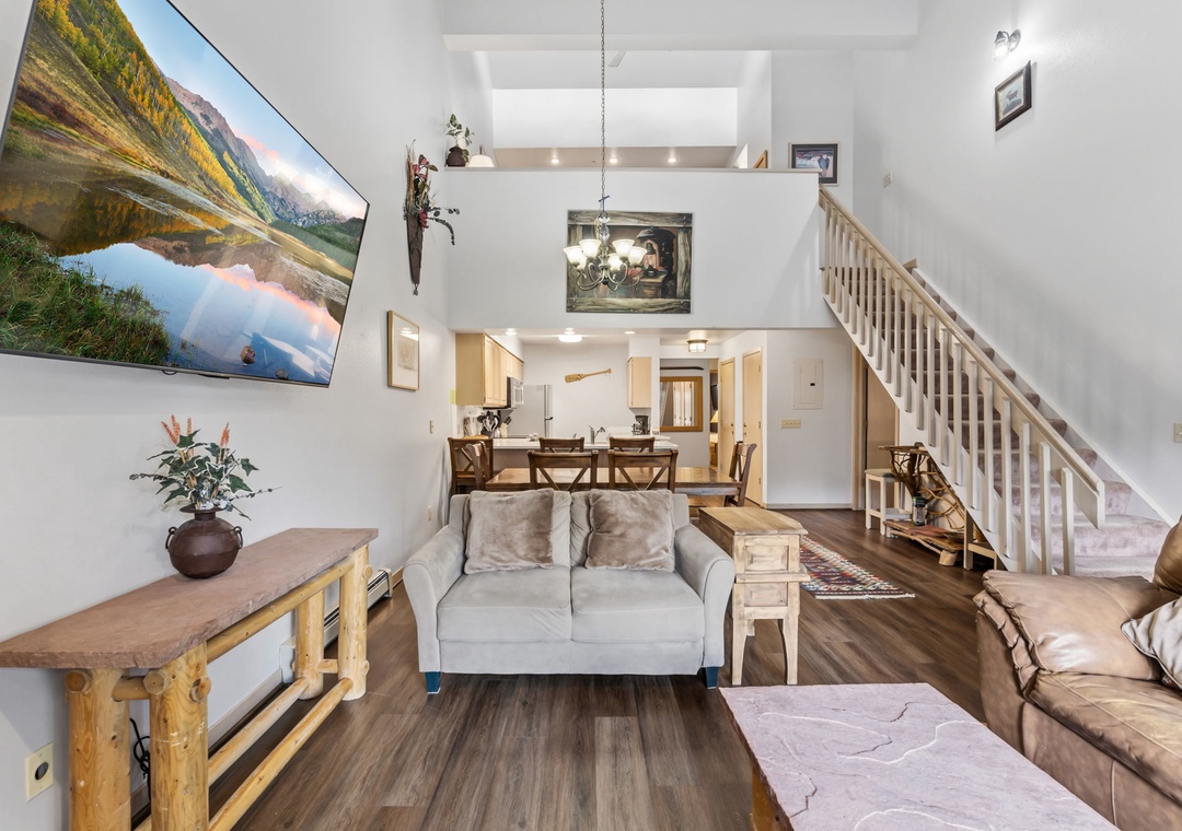 Bright open living space invites you to relax on cozy sofas while enjoying soaring ceilings and natural light streaming through the home. The dining area and loft above create a welcoming, airy atmosphere perfect for your mountain getaway.