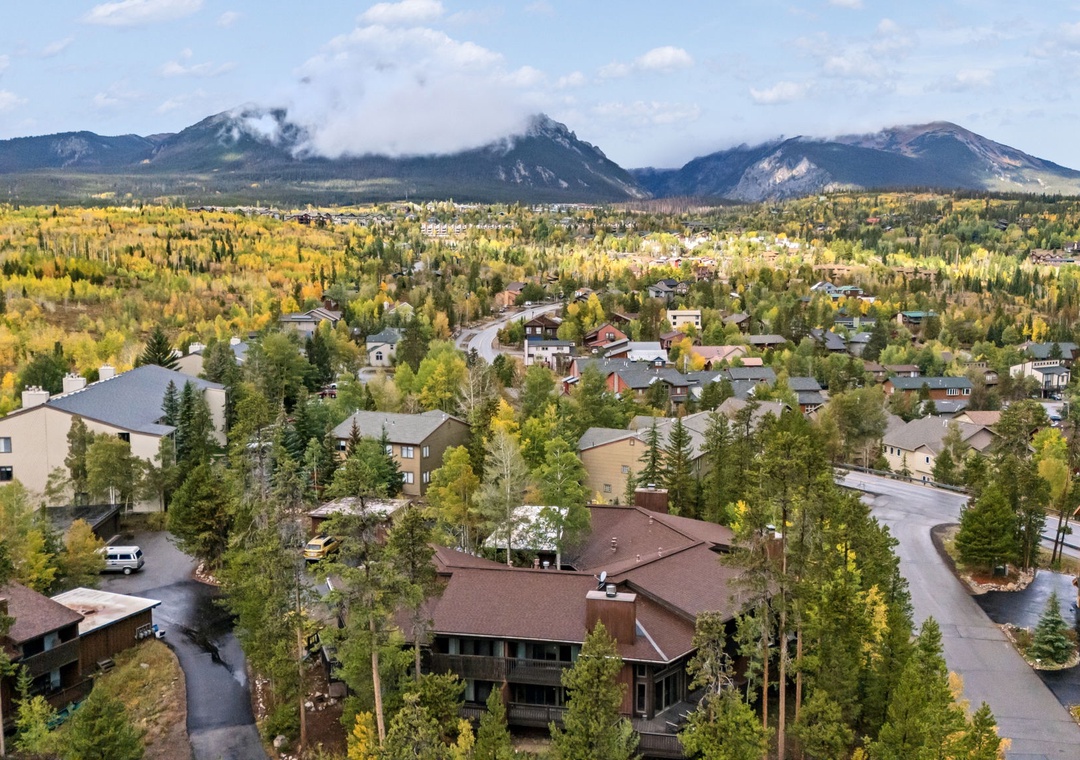 Aerial view of a mountain community nestled among vibrant autumn foliage, with dramatic peaks rising in the distance under a partly cloudy sky.