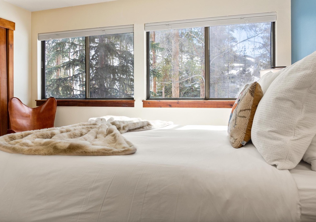 Sink into this plush bed draped in soft linens and a cozy throw, while large windows frame peaceful forest views that make your mountain retreat unforgettable.