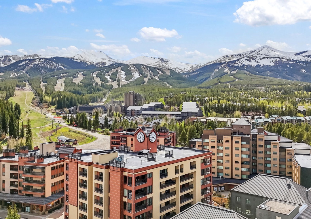 A mountain resort town nestled in a stunning alpine valley, surrounded by snow-capped peaks and evergreen forests, with ski slopes and nearby properties visible across the landscape.