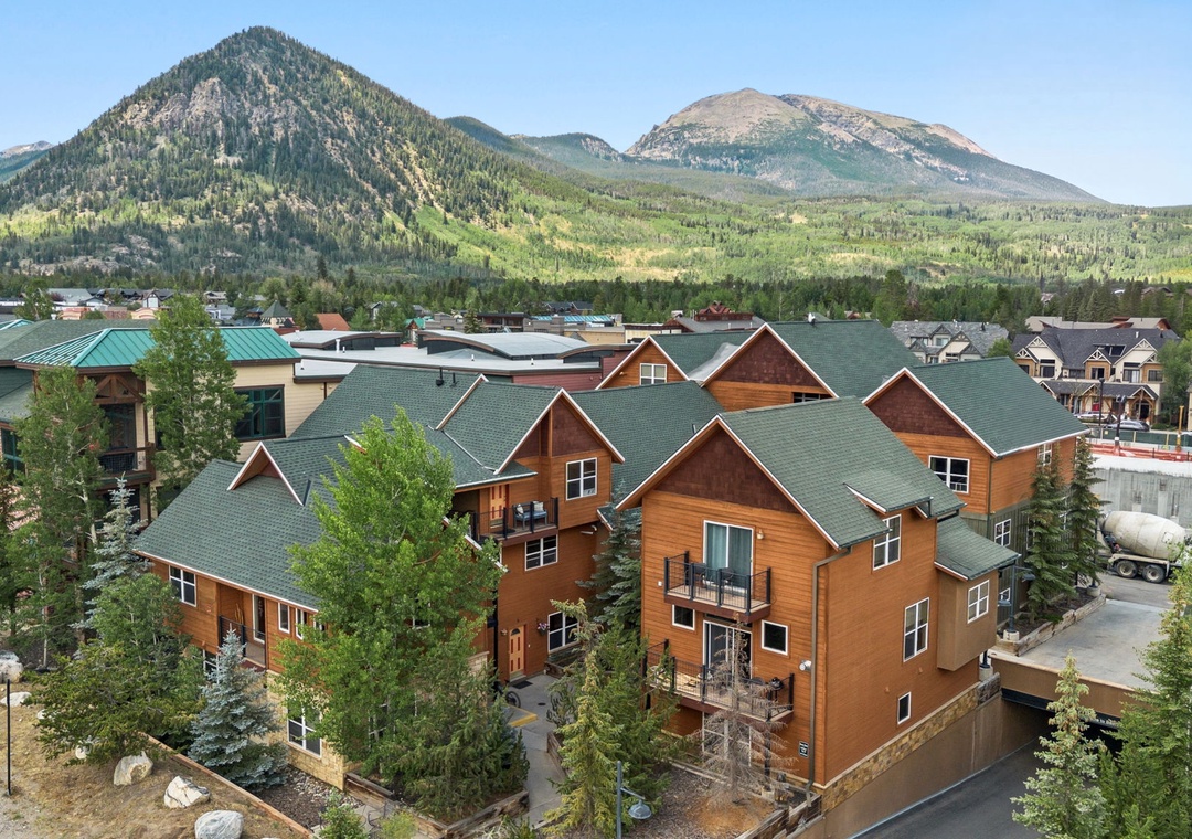 Elevated view of a vacation rental complex nestled in a scenic mountain town, surrounded by forested peaks and residential neighborhoods under clear blue skies.