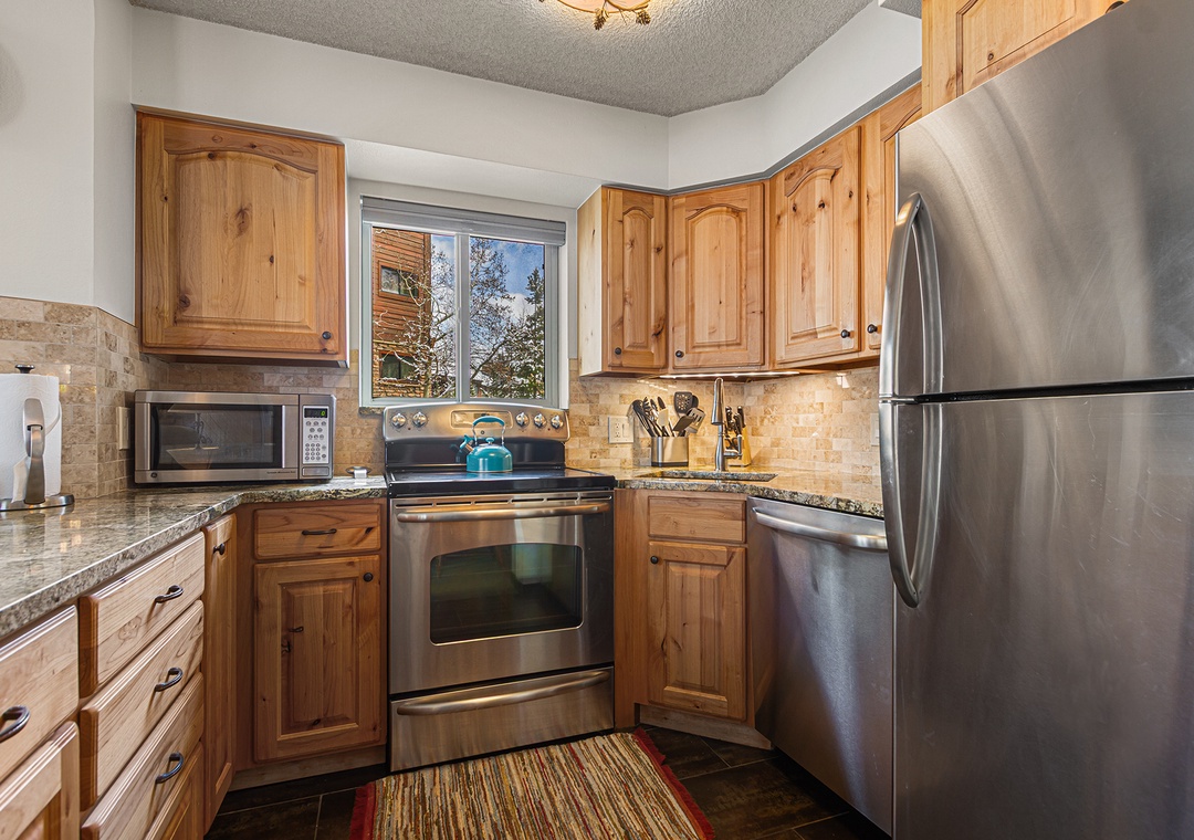 Your fully equipped kitchen awaits with warm wood cabinetry, stainless steel appliances, granite countertops, and mountain views through the window—perfect for preparing home-cooked meals during your stay.